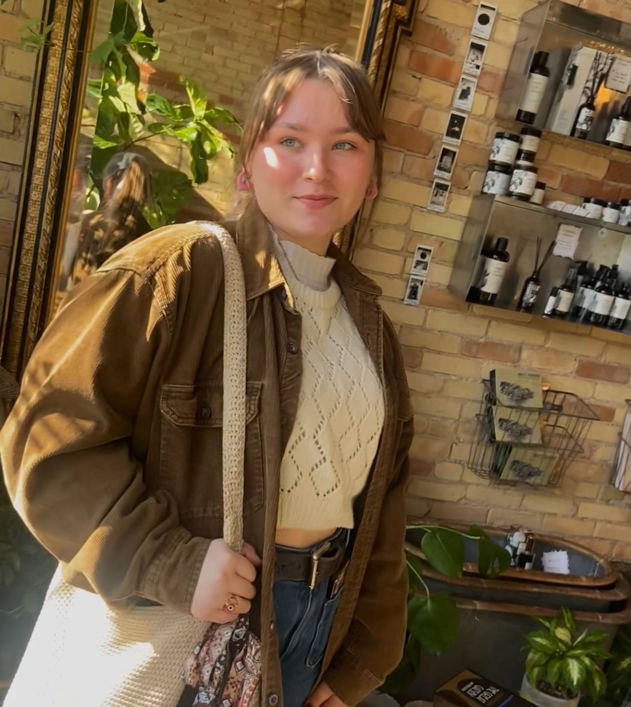 Woman wearing brown jacket and holding cream colored bag standing in store with brick background